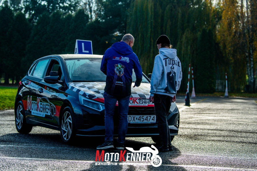 Na zdjęciu widać dwie osoby. Jedną jest instruktor nauki jazdy prowadzący szkolenie na prawo jazdy kategorii B. Druga osoba to kursantka. Osoby stoją przed maską samochodu Hyundai I20. Pojazd ma charakterystyczne, czerwone, szare i czarne malowanie reklamujące szkołę jazdy Hatchback to taki sam pojazd na jakim zdaje się egzaminy w DORD Wrocław