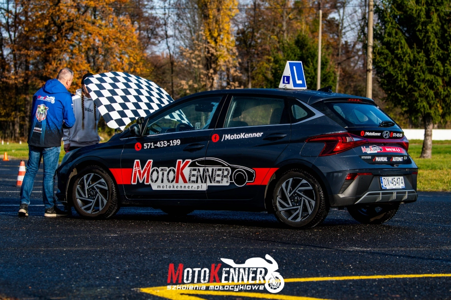 Na zdjęciu uwieczniono samoch&oacute;d szkoleniowy z czerwono-szarymi akcentami i napisami na bokach wyraźne oznaczenia szkoły jazdy oraz numery kontaktowe, co podkreśla ich przeznaczenie do nauki jazdy. Ujęcie to ilustruje ważny aspekt kursu prawa jazdy kategorii B, kt&oacute;ry obejmuje naukę prawidłową eksploatacje pojazdu i dbanie o środowisko naturalne