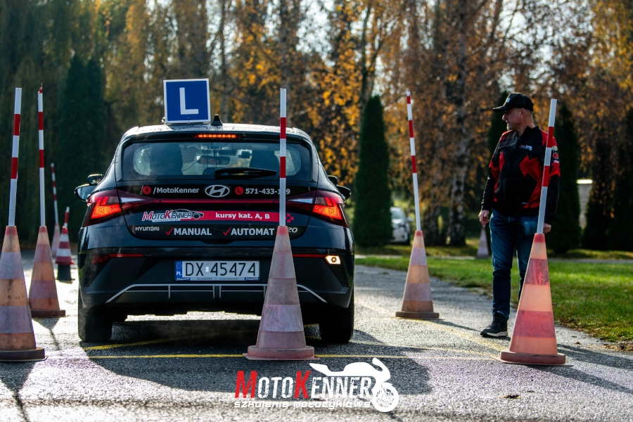 Fotografia przedstawia samoch&oacute;d nauki jazdy podczas wykonywania zadania egzaminacyjnego jazda po łuku. Fachowe określenie dla tego zadania to jazda pasem ruchu do przodu i do tyłu. Zadanie wykonywane jest na placu manewrowym. Pojazd ma tylne światła stopu włączone. Na jego tylnej klapie widnieje napis z danymi kontaktowymi do szkoły nauki jazdy MotoKenner