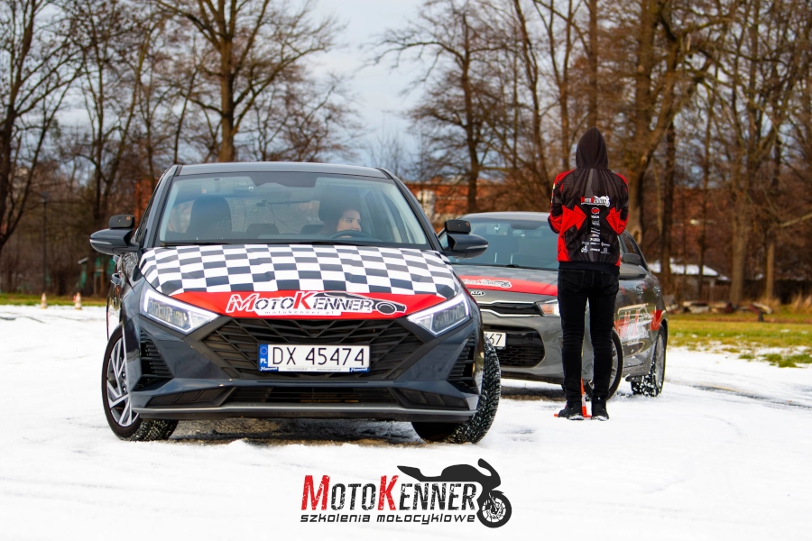 Na zdjęciu przedstawiono dwa samochody kursu prawa jazdy kategorii B w trakcie jazdy podczas specjalnego szkolenia zimowego. Pierwszy to ciemnoszary hatchback z czerwoną maską i bocznymi napisami 