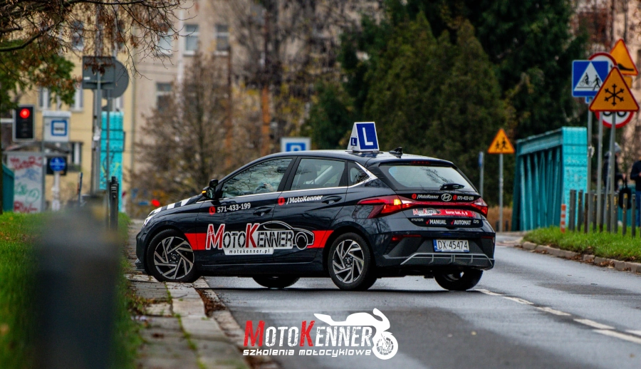 Na fotografii przedstawiono samoch&oacute;d skręcający w lewo przed mostem nad rzeką.  Na boku auta widnieje napis z charakterystycznym logo firmy szkoleniowej